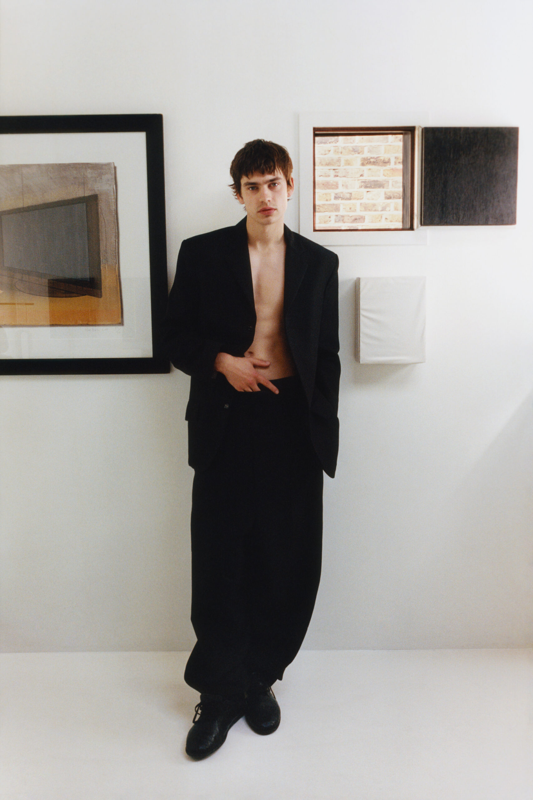 Spike Fearn wears a black suit jacket and trousers and leans against a white wall with picture frames