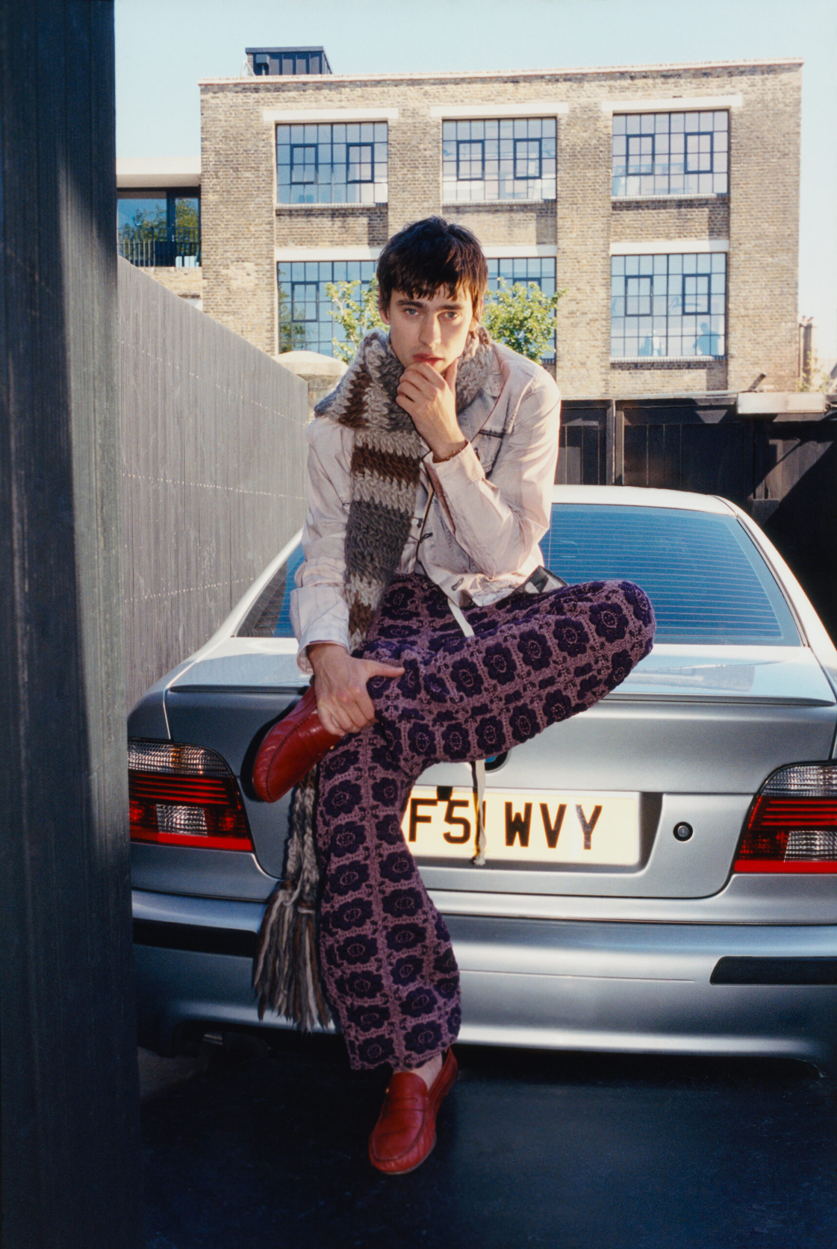 Spike Fearn sits on the boot of a silver sedan with his finger touching his lip and his hand clutching his ankle.