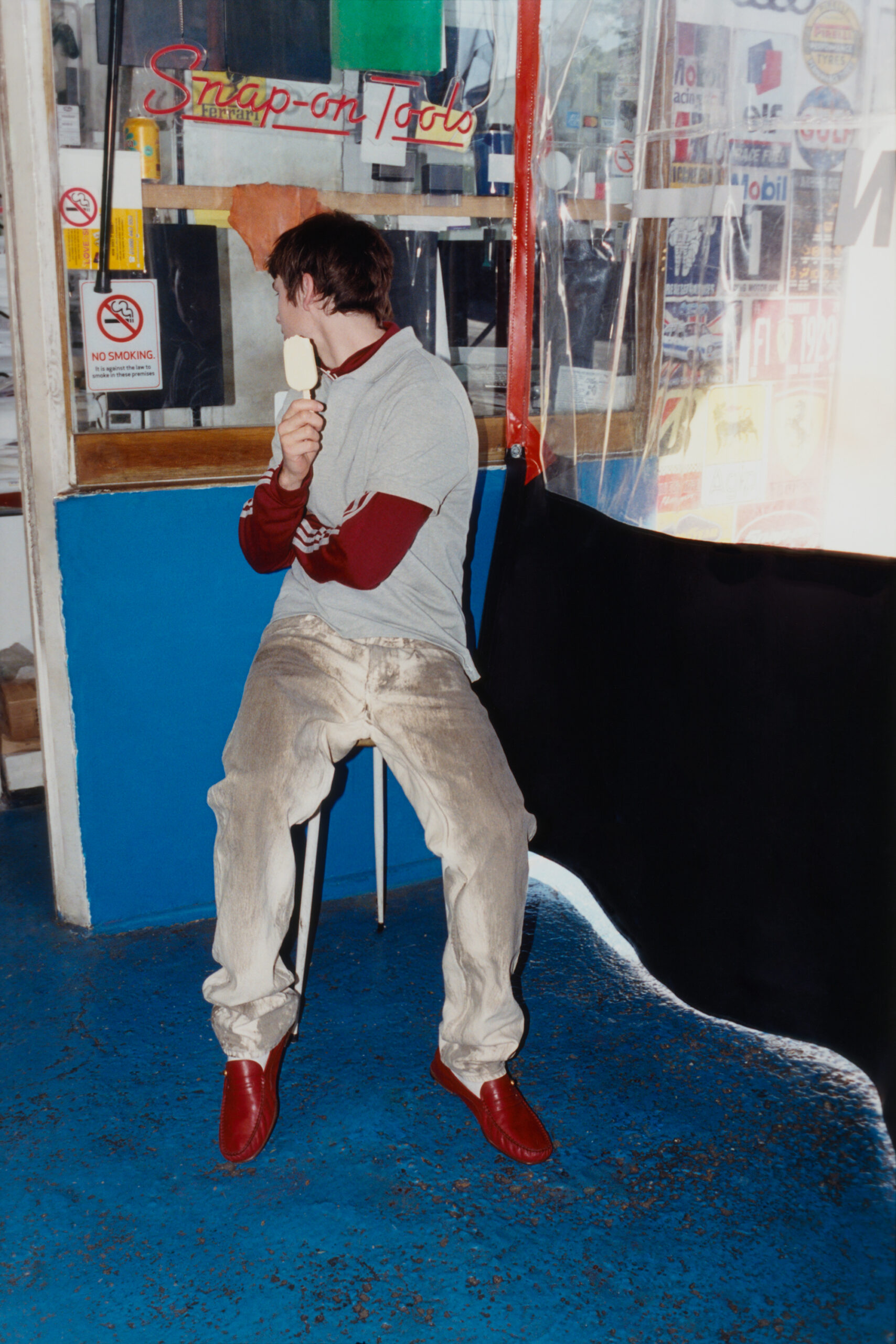 Looks away from the camera holding a popsicle while sitting on a stool.
