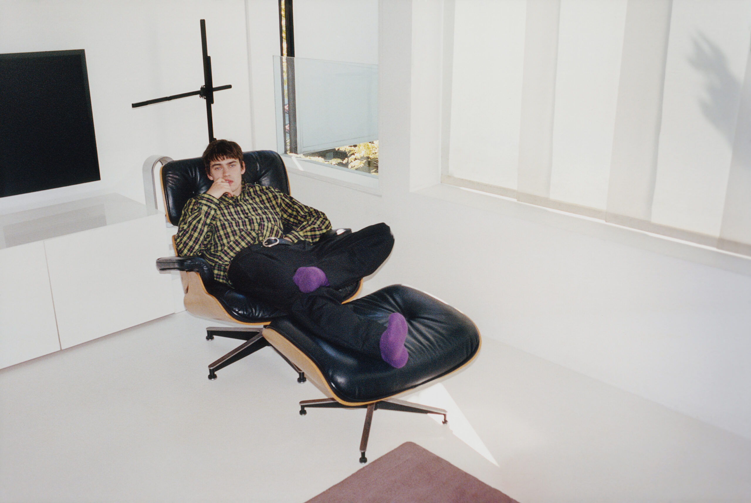 Lays back on a reclining leather office chair with one foot propped on the opposite knee.