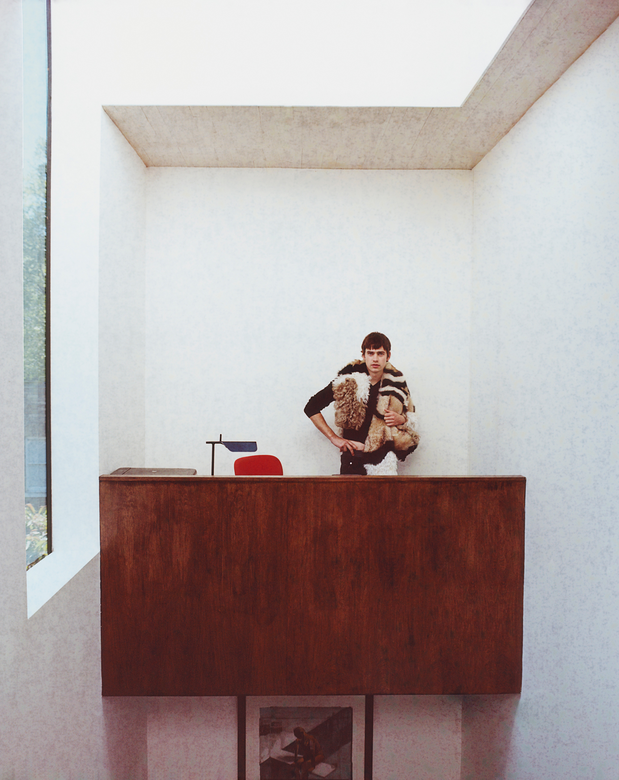 Stands behind a large wooden desk with one hand on his hip as a fur scarf is draped over his neck.