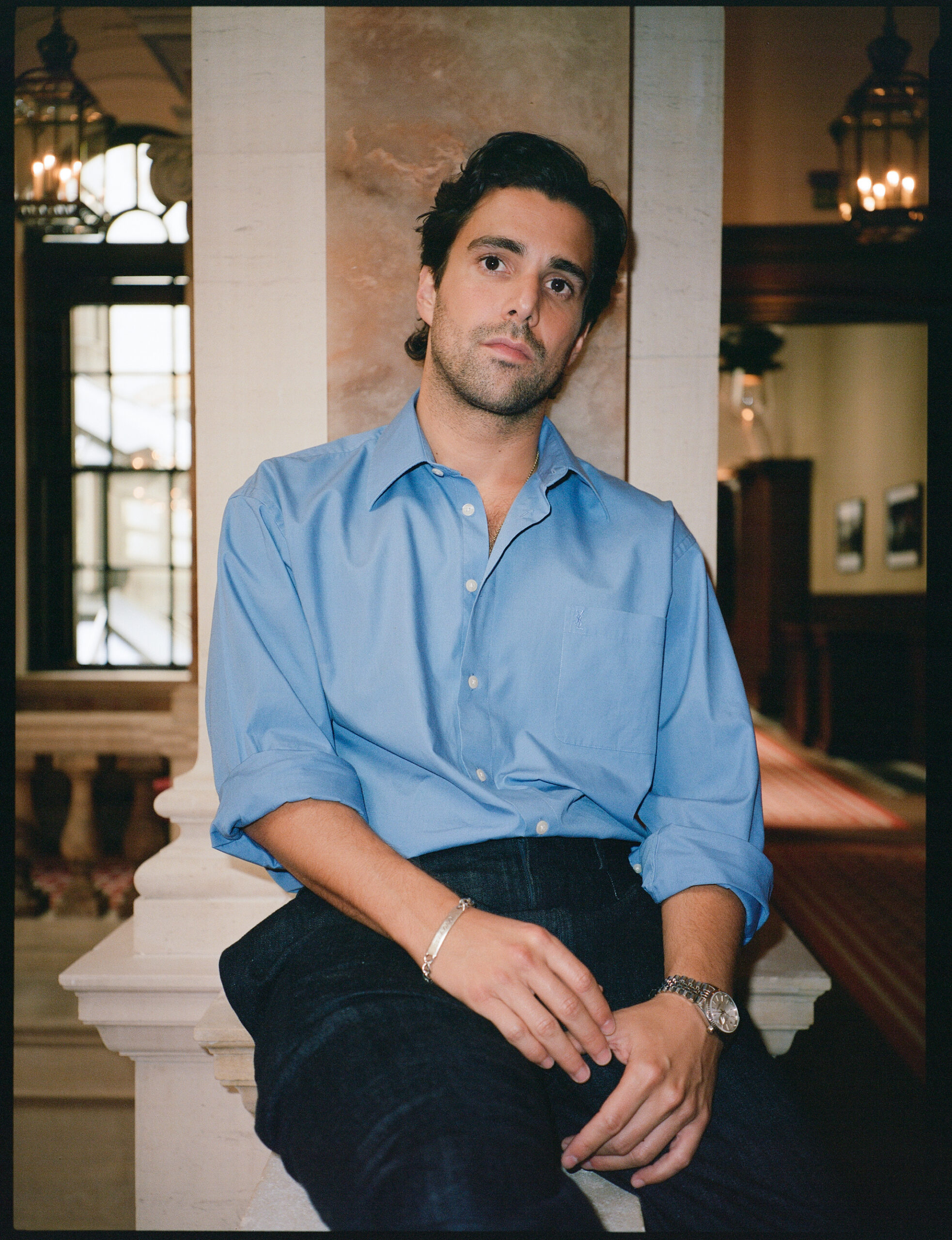 Fabian Frankel wearing a blue button up and black pants leaning against a white column