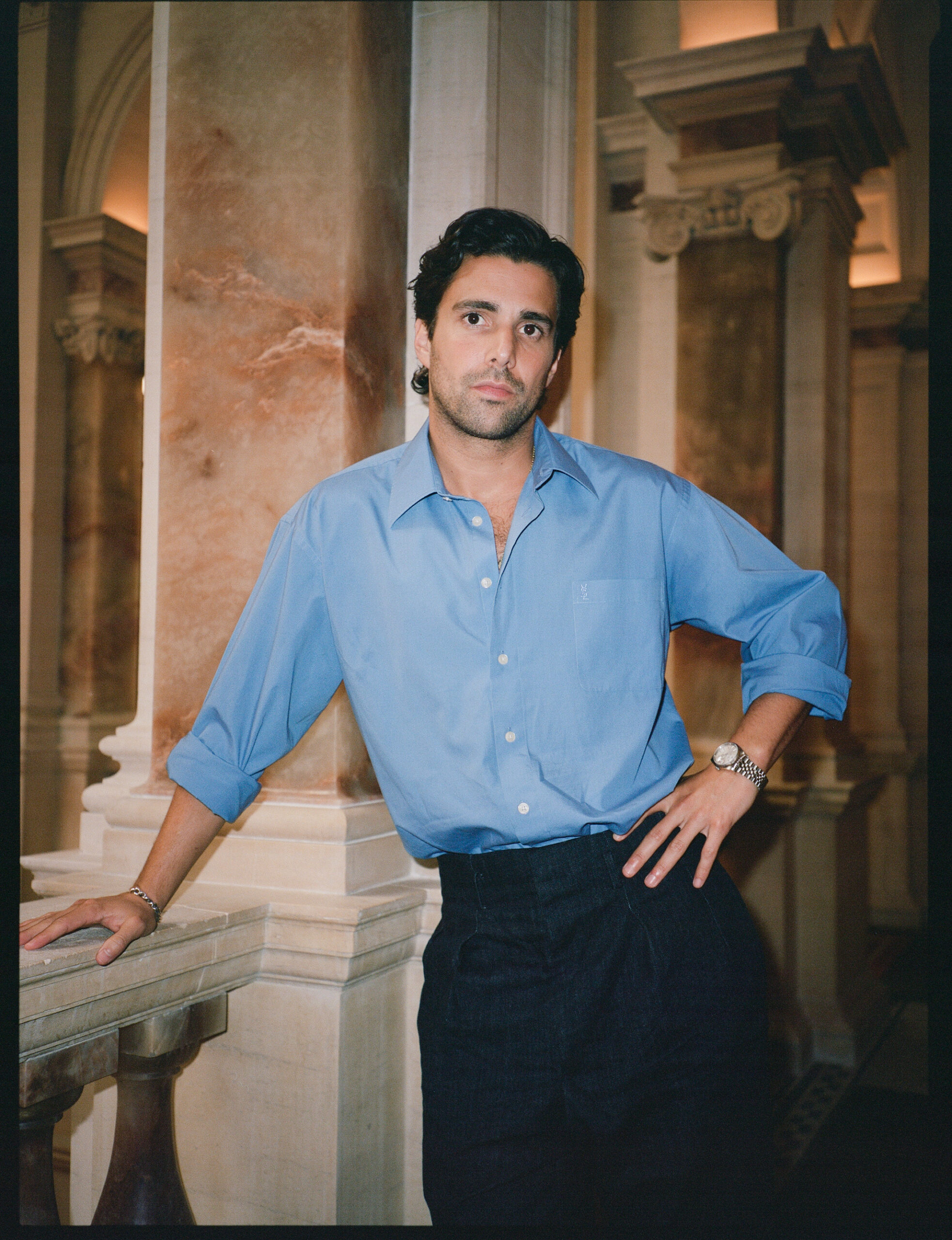 Fabian Frankel with his left hand on his left hip and his right hand leaning against a white stone railing, wearing a blue long sleeve button up and black trousers