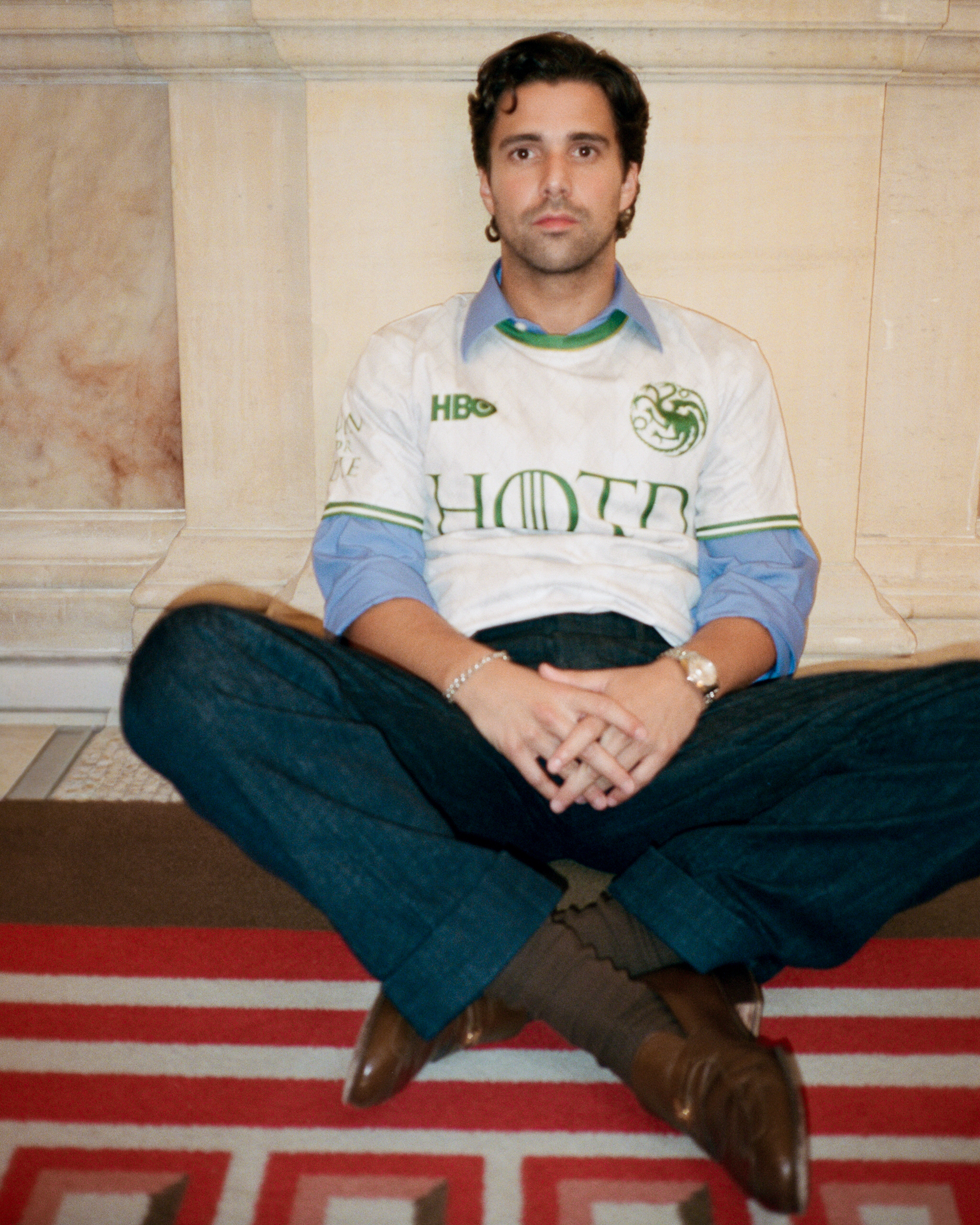 Fabian Frankel sitting cross legged on a red carpet, wearing blue trousers, brown shoes, and a white and green jersey over a blue button up