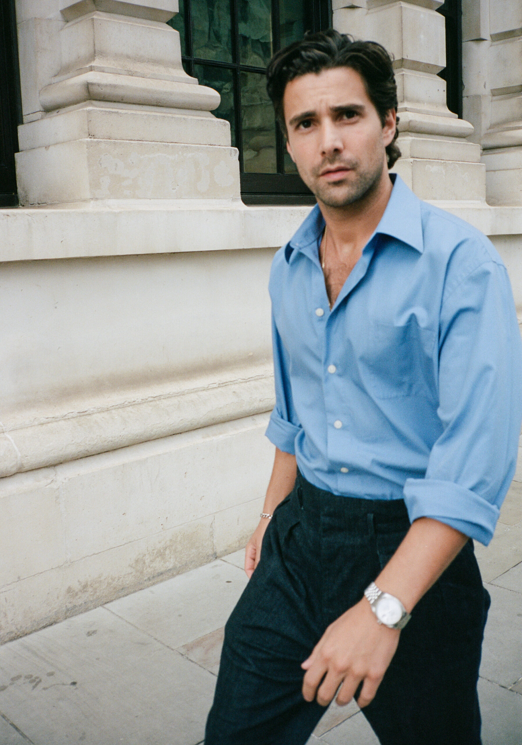 Fabian Frankel wearing a blue long sleeve button up collared shirt, black trousers, and a shiny silver watch, walking by a white building and turning his head to look at the camera