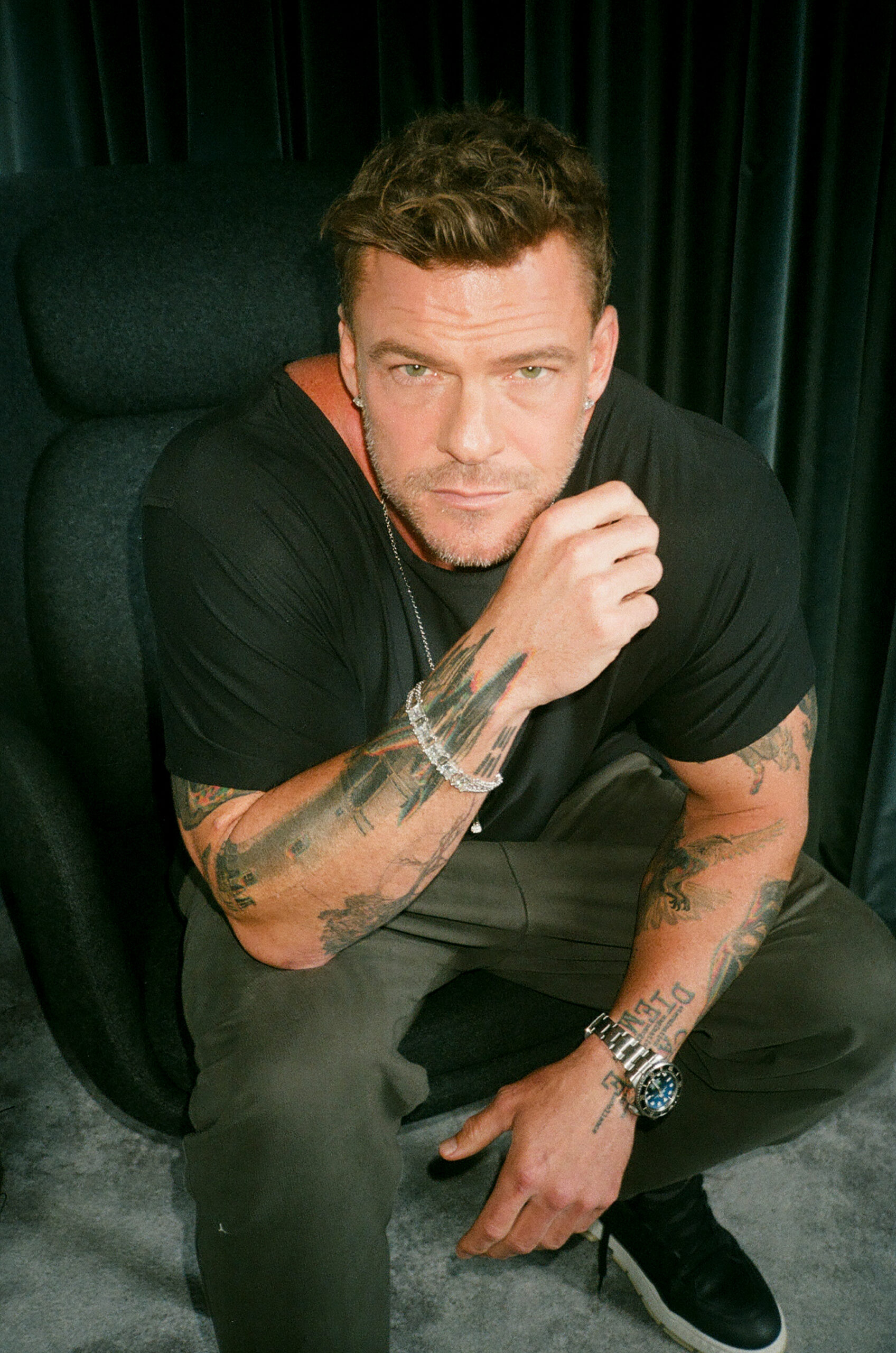 Alan Ritchson sits down on chair dressed in black and stares at camera.