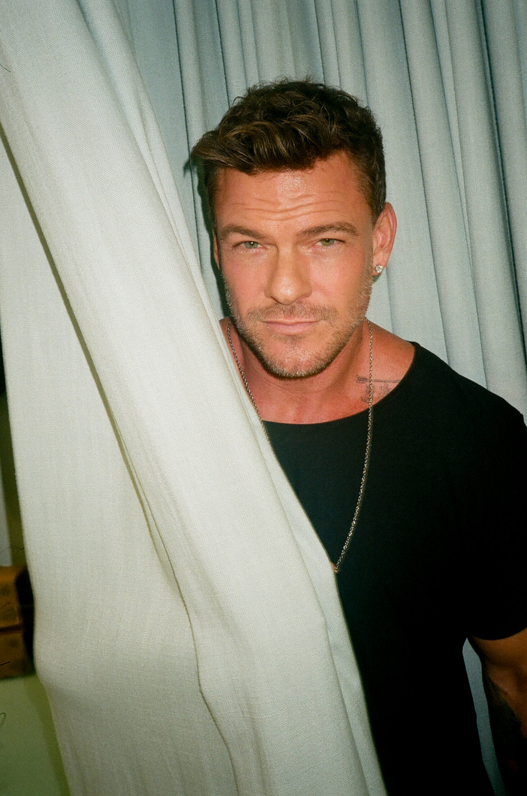 Alan Ritchson stands behind curtain and stares at camera. 