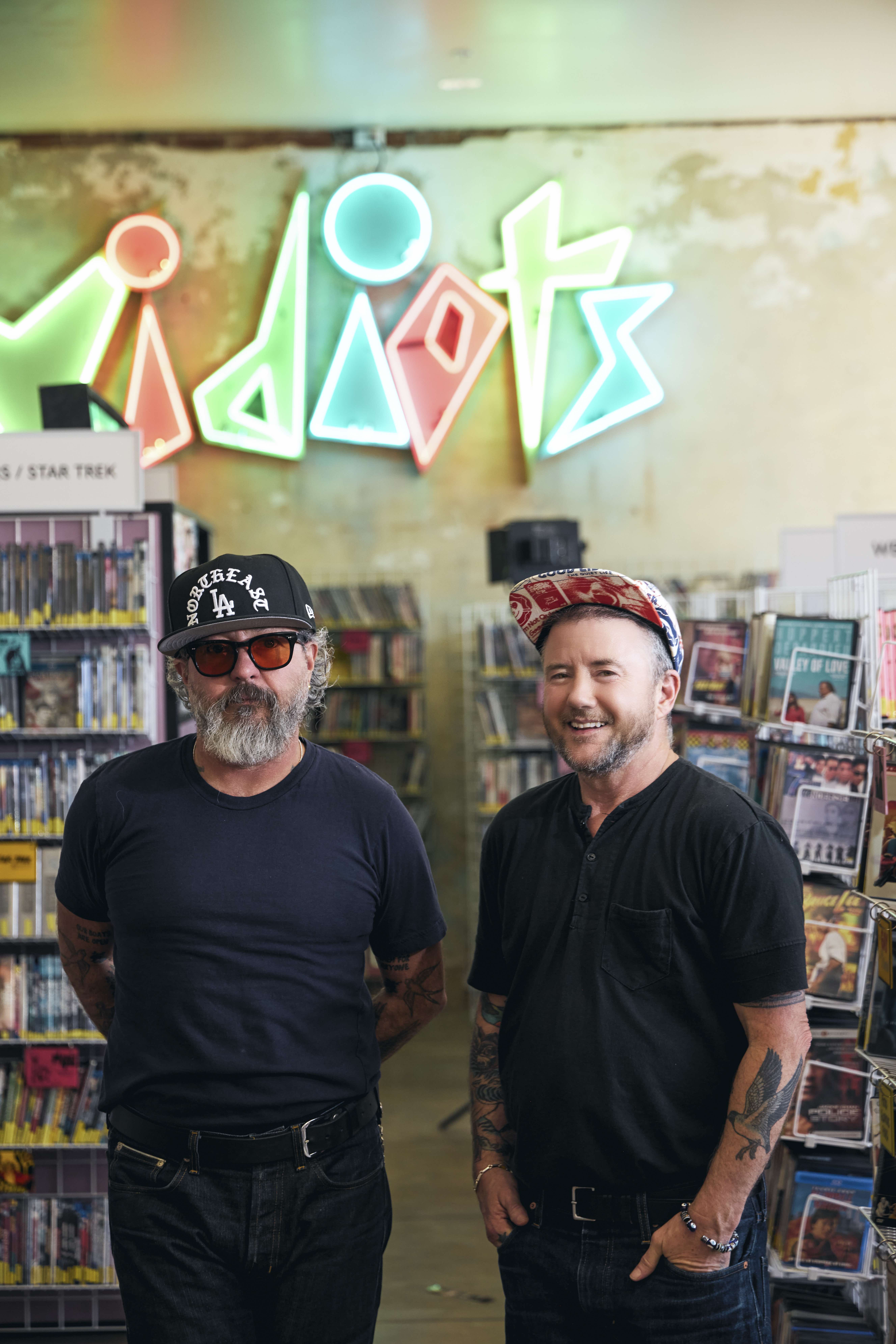 Harry Dodge and Silas Howard stand together in a colordul DVD store smiling at the camera