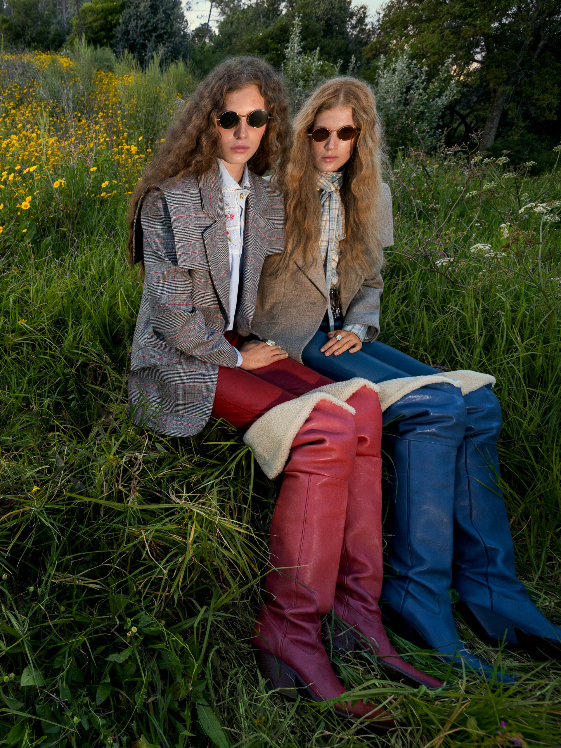 Two models sit next to each other in a field of grass in blazers, sunglasses, and leather boots.