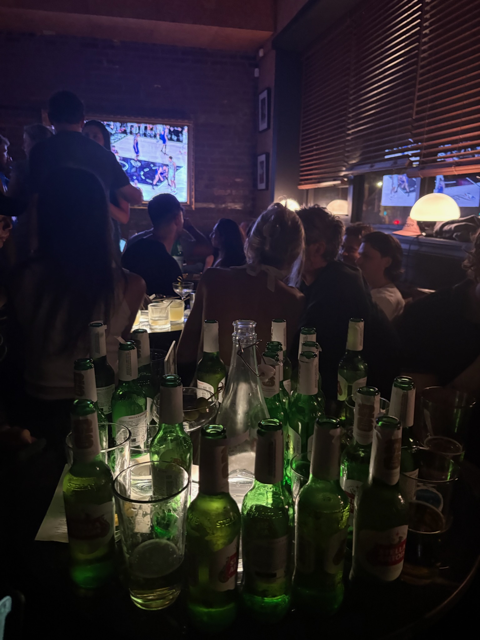Several bottles sit accumulated at the table at a bar. The Knicks game plays on TV in the background.