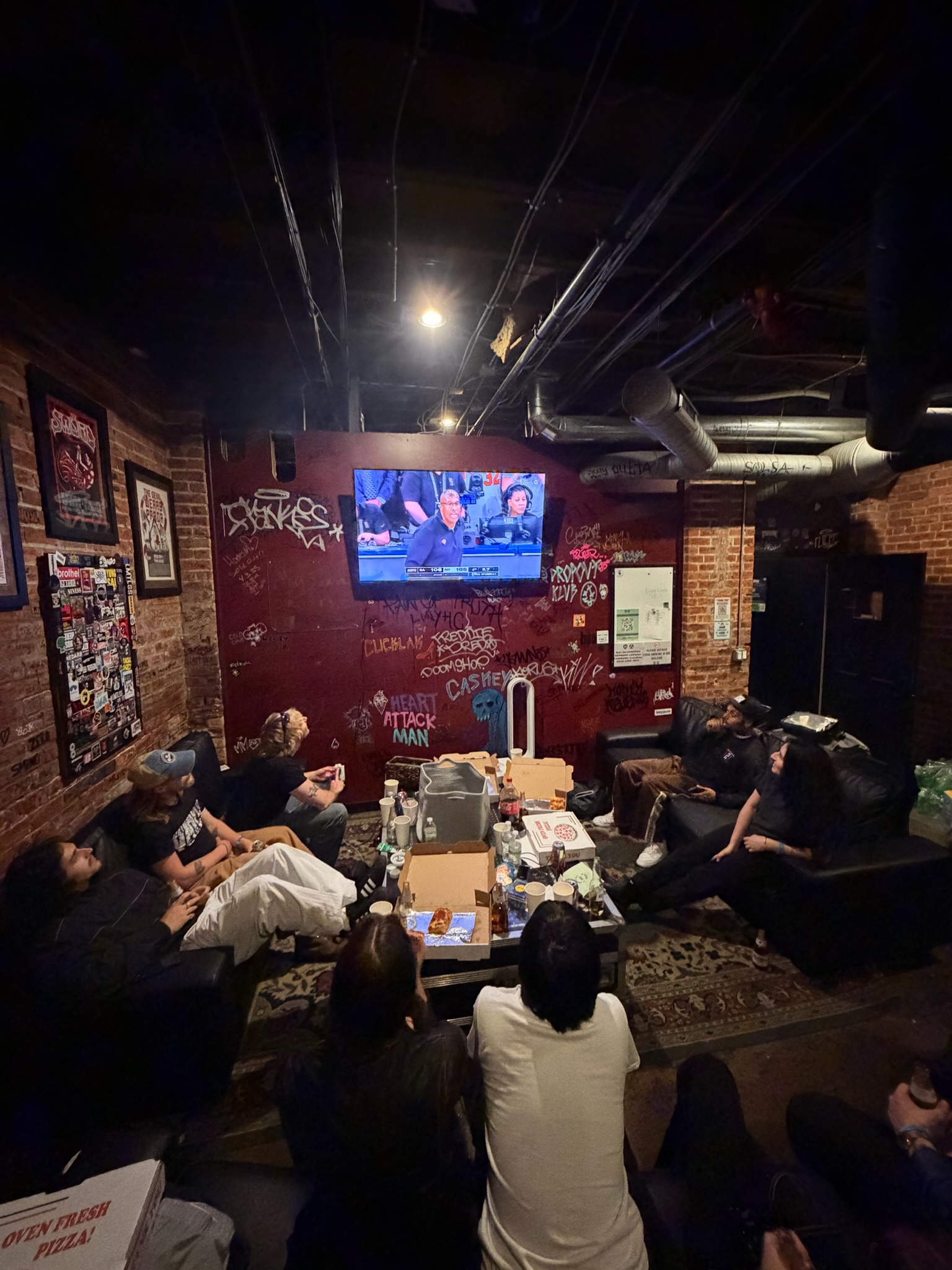 Members of Villanelle and their roadies sit on couches watching the Knicks game on TV.