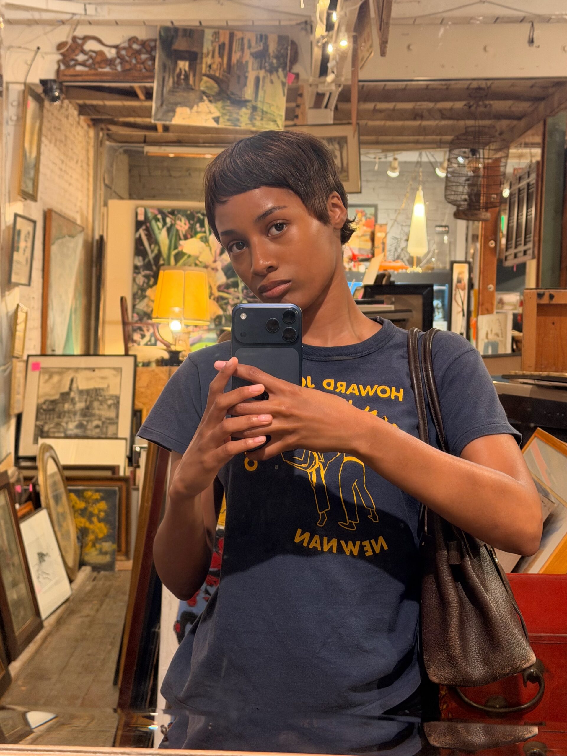 A mirror selfie of girlsweetvoiced with paintings in the back, dressed in a blue shirt with a leather bag on her shoulder.