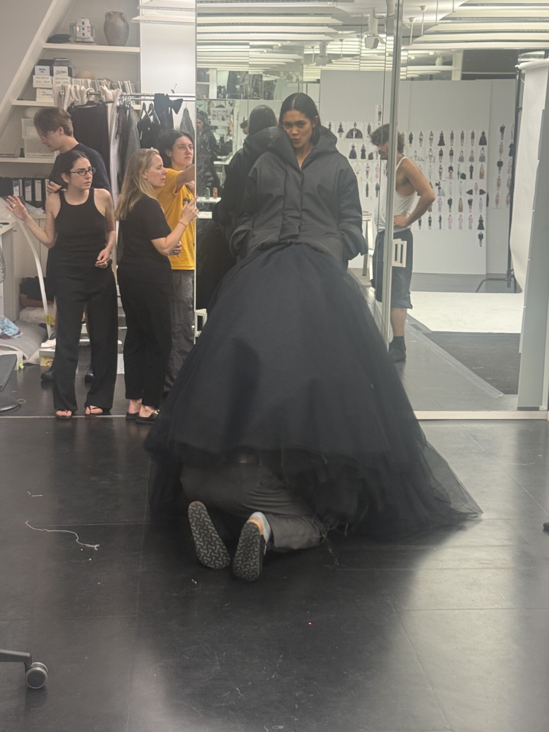 A model backstage at Duran Lantink's show wearing a black dress with black tool in the front. Someone has climbed underneath the tool skirt to adjust the look
