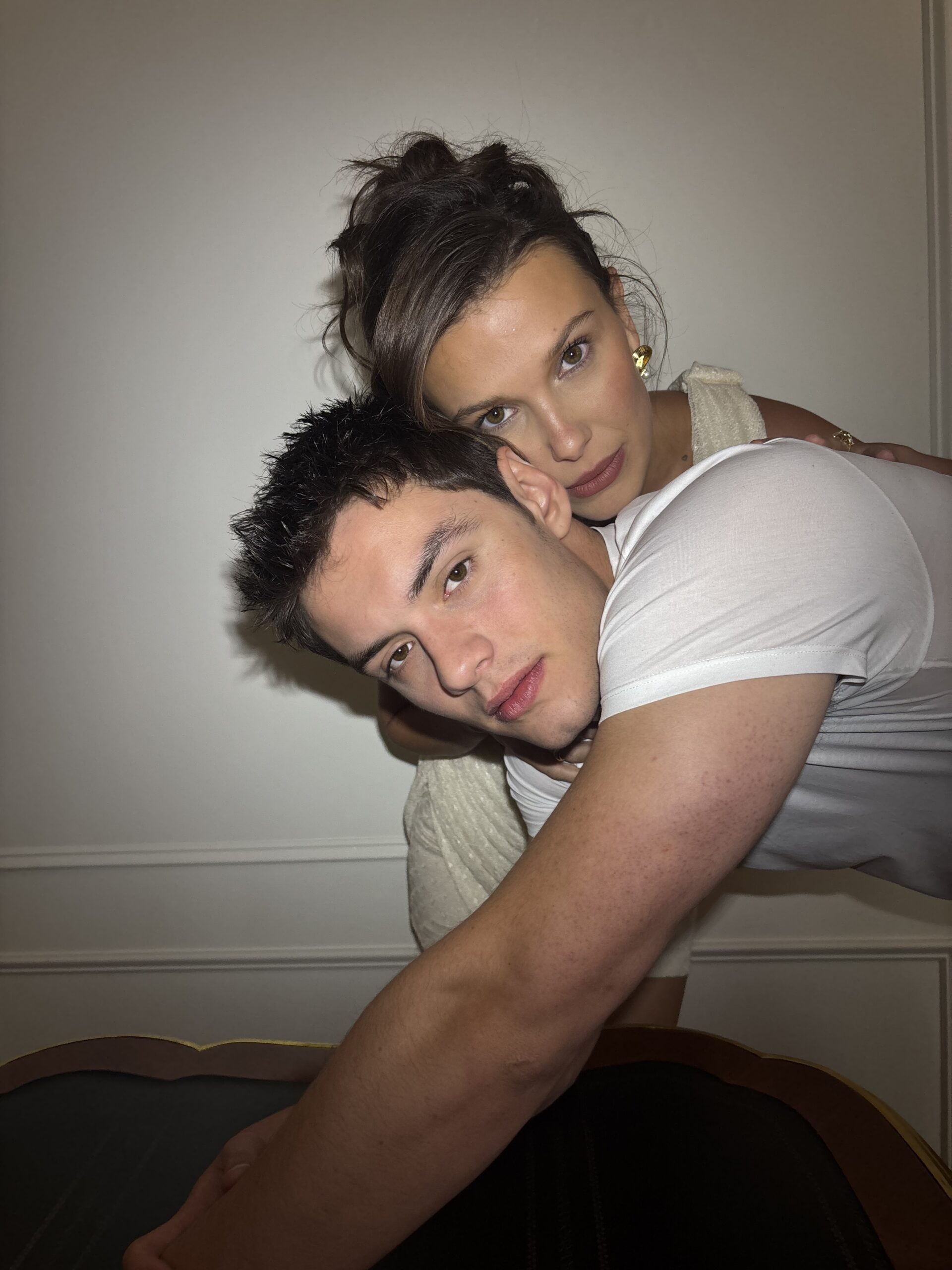 Millie Bobby Brown and Louis Partridge lean over the wooden bench. Louis Partridge wears a white t-shirt. Brown stands behind him resting her head on his.