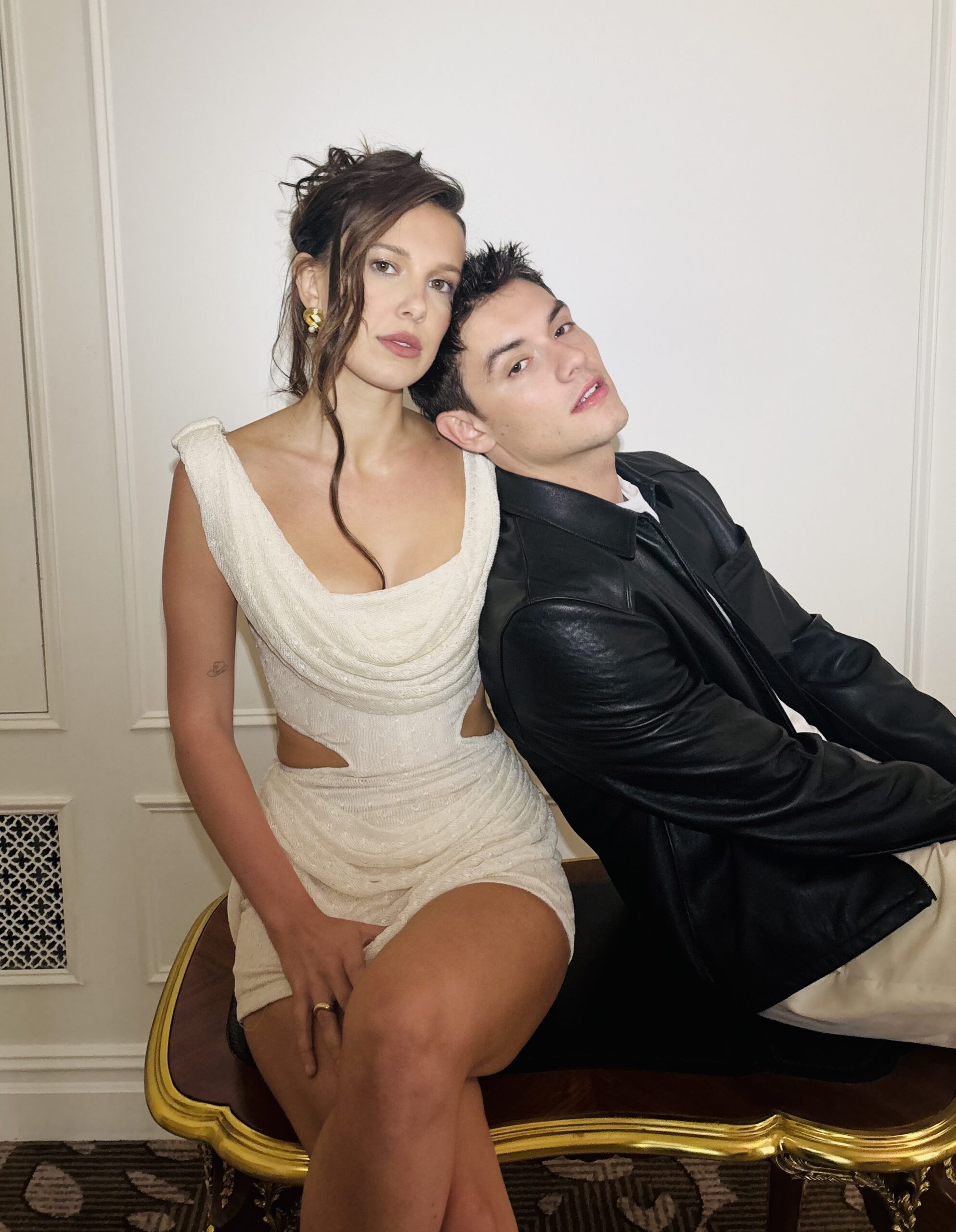Millie Bobby Brown and Louis Patridge sit on a polished wooden bench. She wears a white cut-out mini dress and he wears a black leather jacket and beige trousers. He rests his head on her shoulder.
